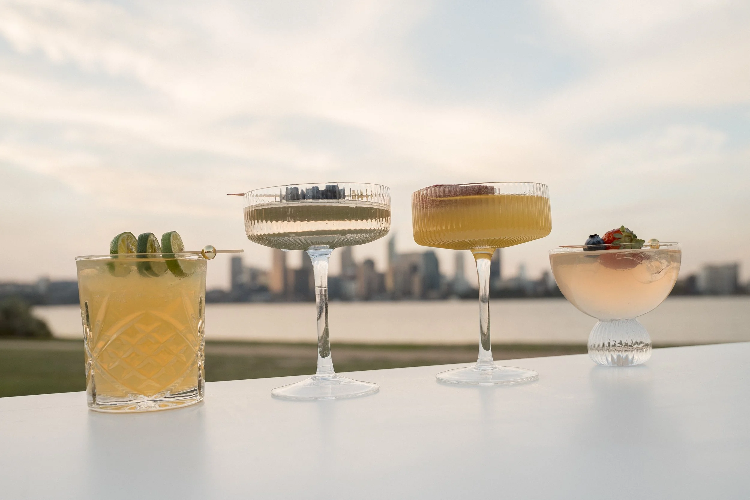 Four cocktails with different garnishes on a table outdoors, against a city skyline and cloudy sky in the background.