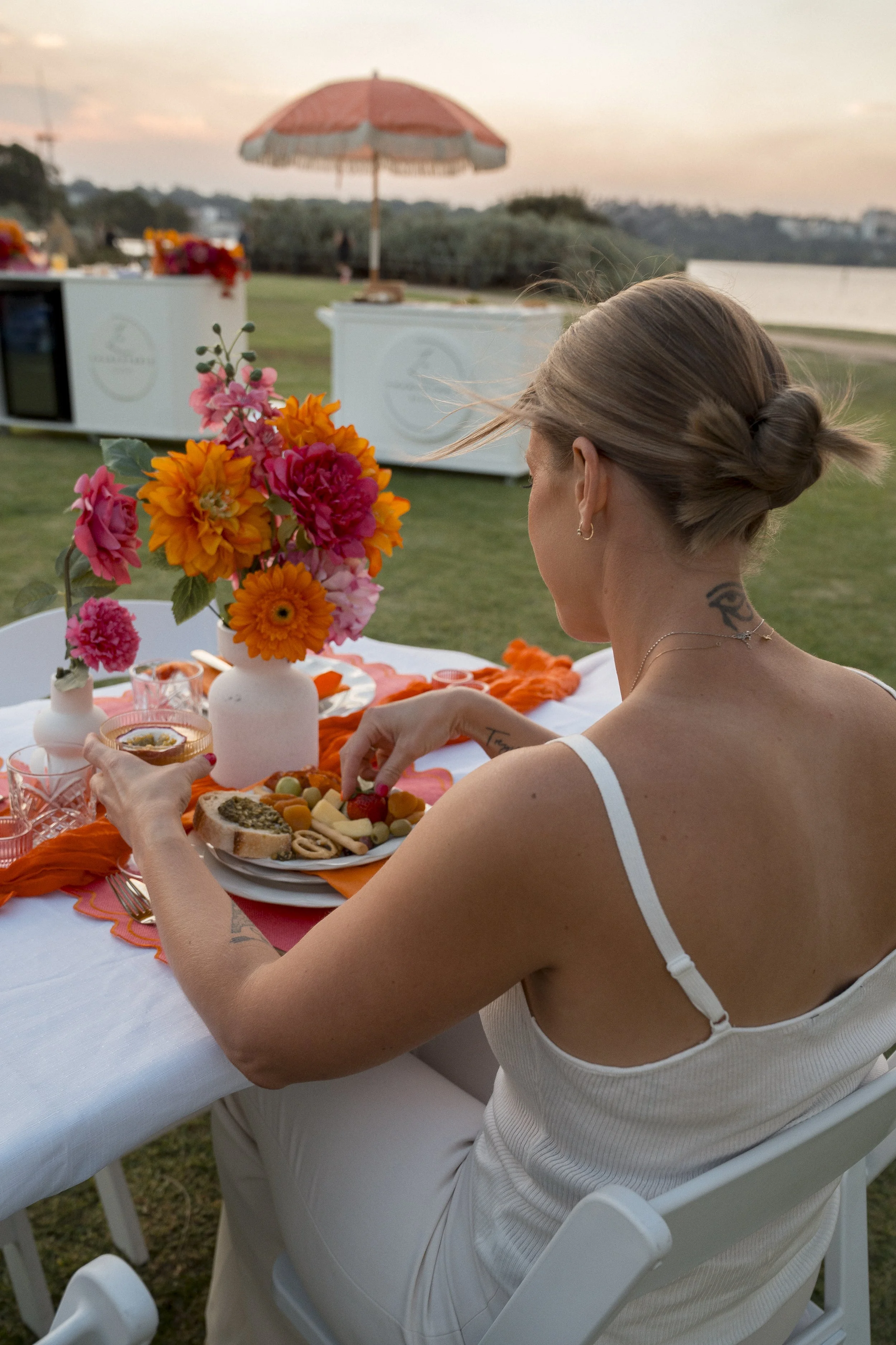 A woman with a tattoo on her arm, wearing a white tank top and white pants, sitting at an outdoor table during sunset, with a lake and shoreline in the background. The table is decorated with a vibrant pink, orange, and red floral arrangement, and it