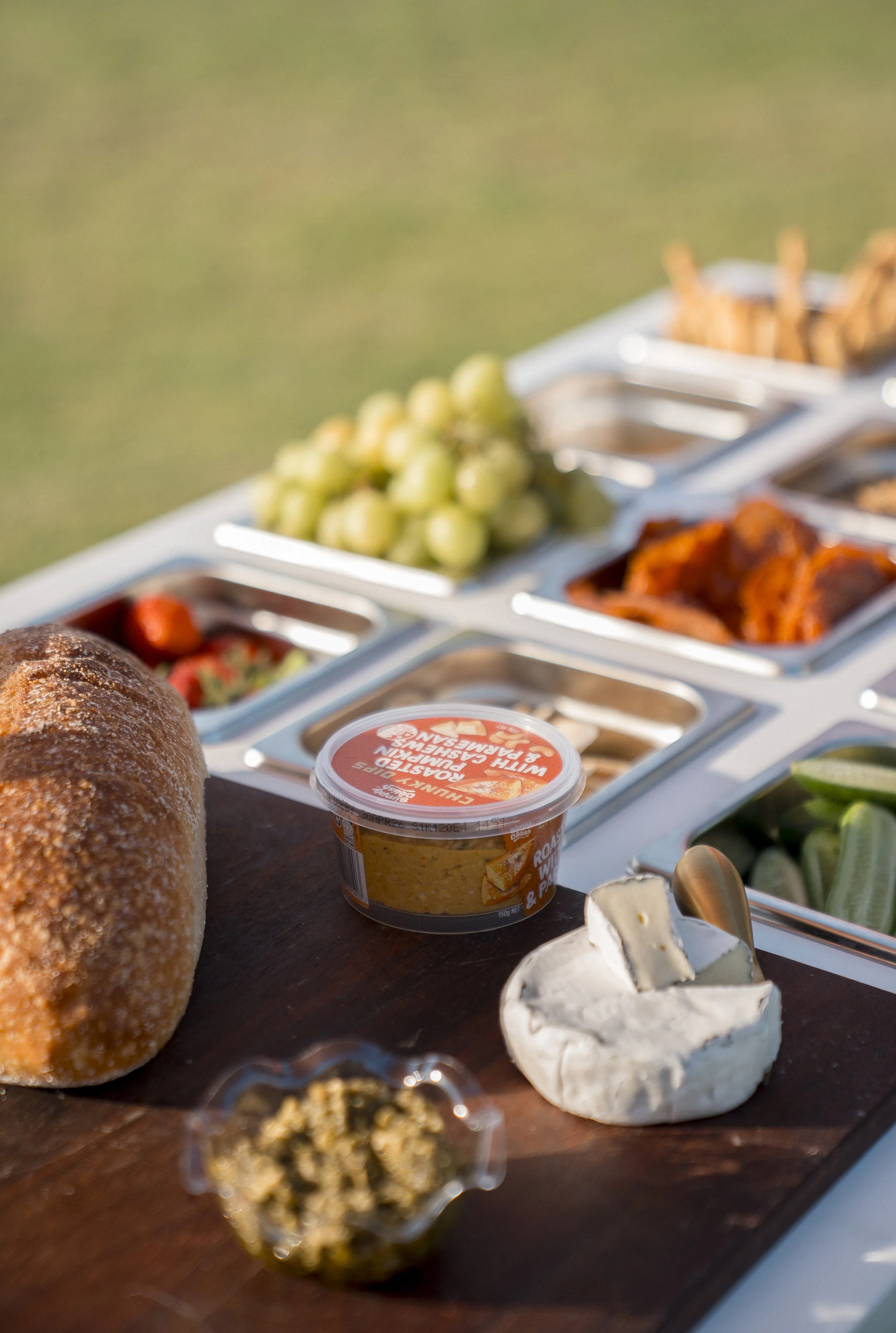 Close-up of a cheese platter with various cheeses, grape clusters, bread roll, and other appetizers on a wooden board, blurred background of a buffet table.