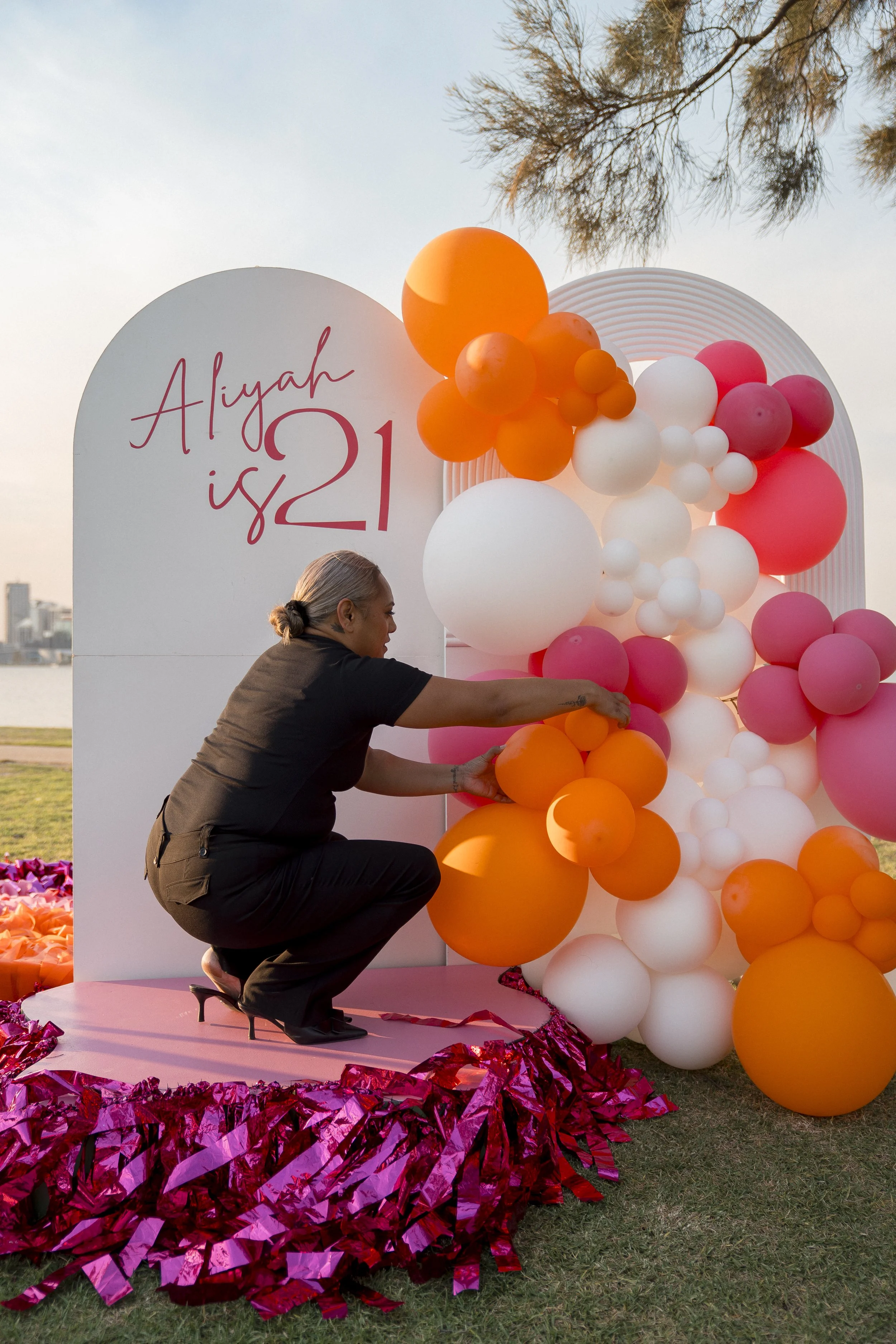 Woman arranging orange, pink, white, and yellow balloons on a pink platform at an outdoor event during sunset, with a city skyline and trees in the background, and a sign that says "Aliyah is 21."