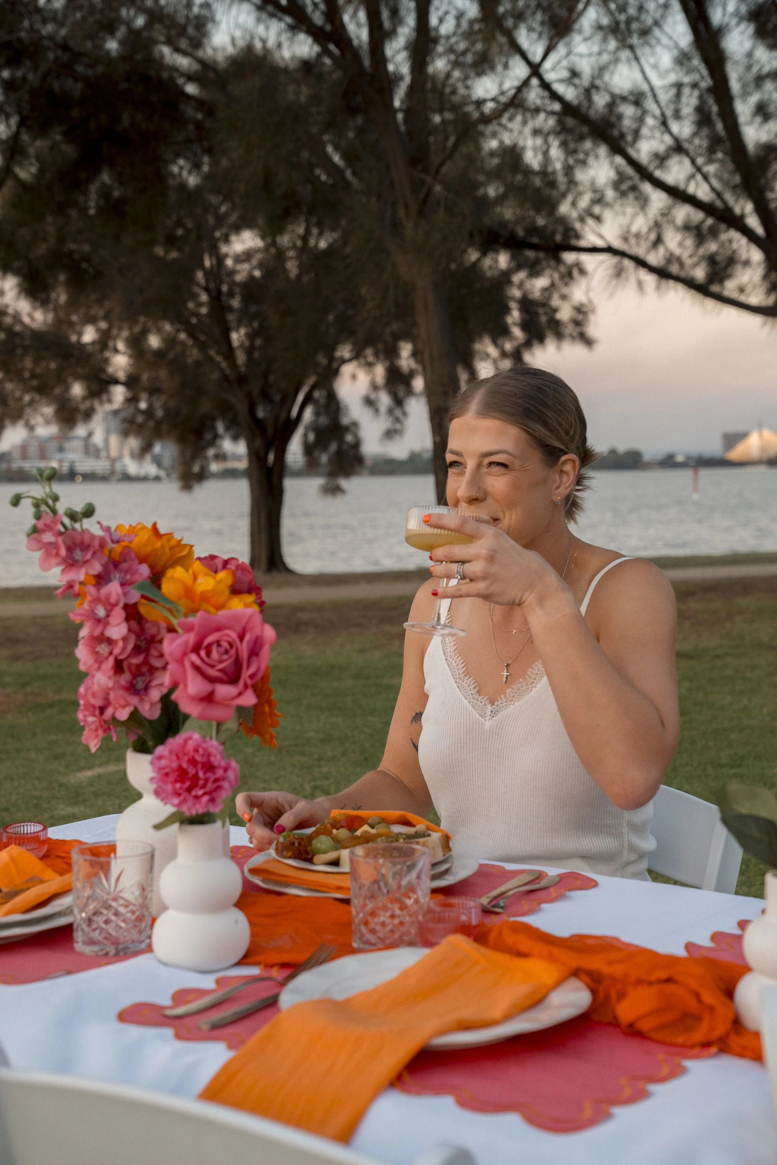 A woman seated at a decorated outdoor table, holding a glass of drink, with a waterfront and trees in the background during sunset.