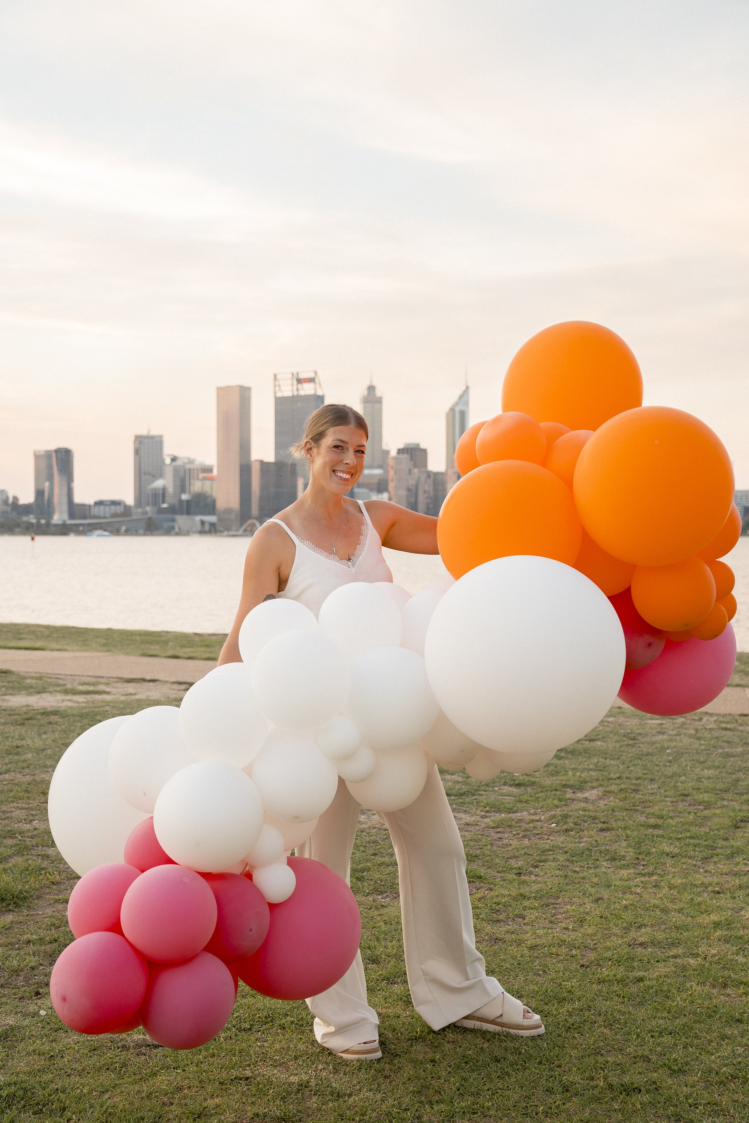 A woman in white pants and a white sleeveless top standing outdoors holding colorful balloons with a city skyline in the background during sunset.