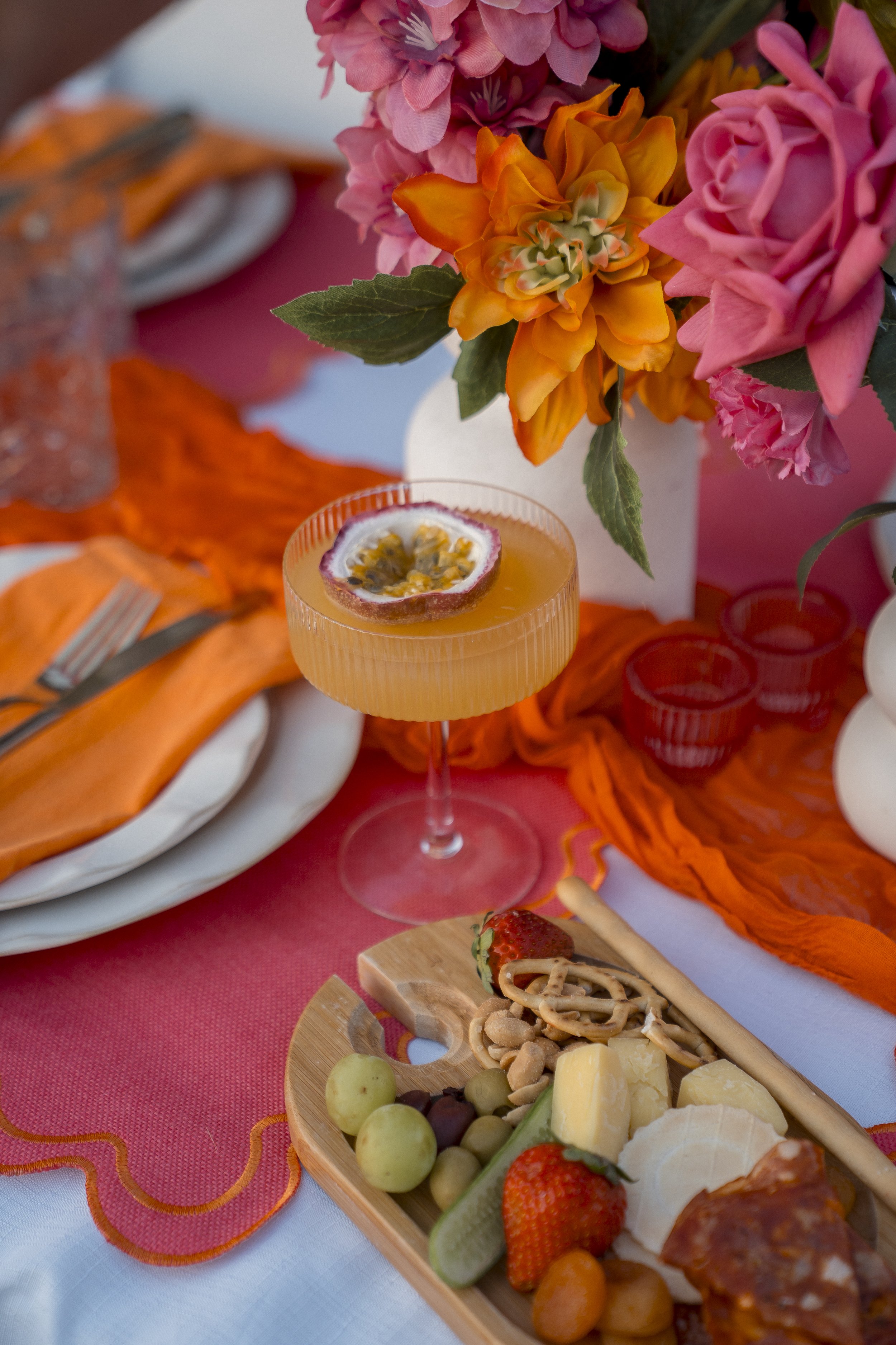 A festive table setting with a colorful bouquet of flowers and an assortment of snacks and beverages, including cheese, strawberries, grapes, crackers, and a passion fruit drink garnished with passion fruit pieces, all arranged on a pink and orange t