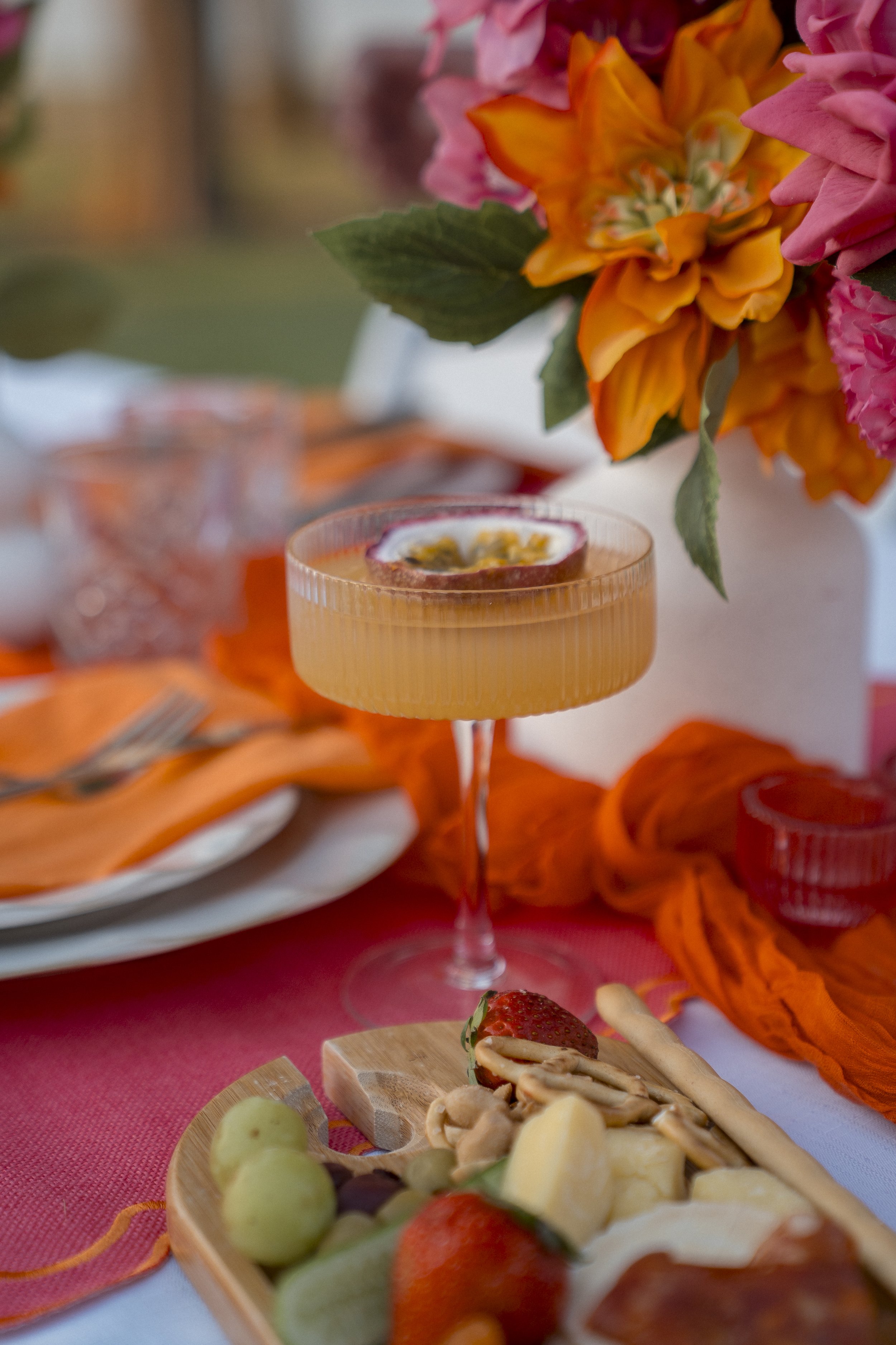 Close-up of a festive table with an orange cocktail in a pink glass, decorated with passion fruit, and a platter of cheese, grapes, strawberries, and nuts, with a bouquet of pink and orange flowers in the background.