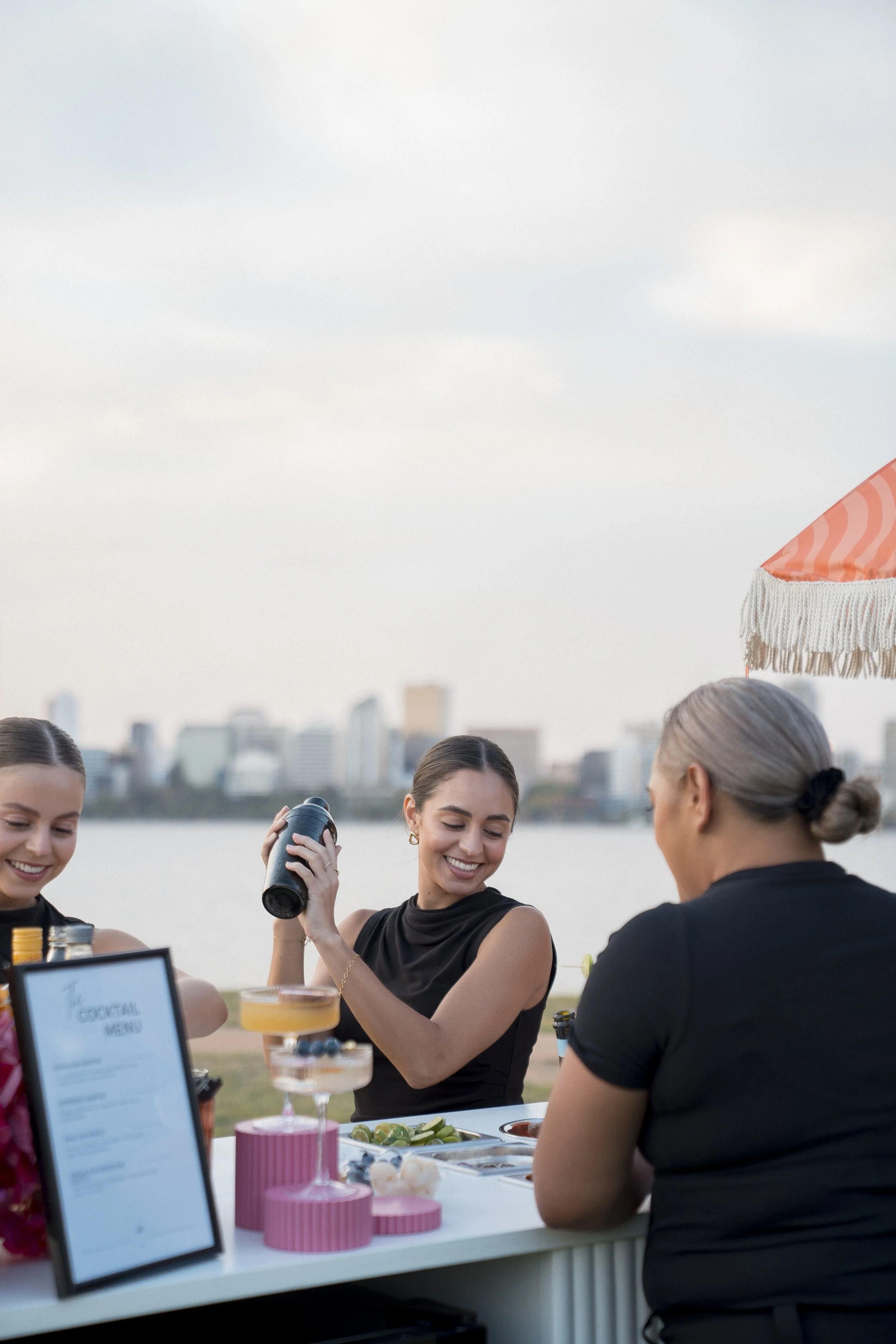 A group of women at an outdoor event, with a city skyline in the background, enjoying drinks and socializing under a large umbrella.
