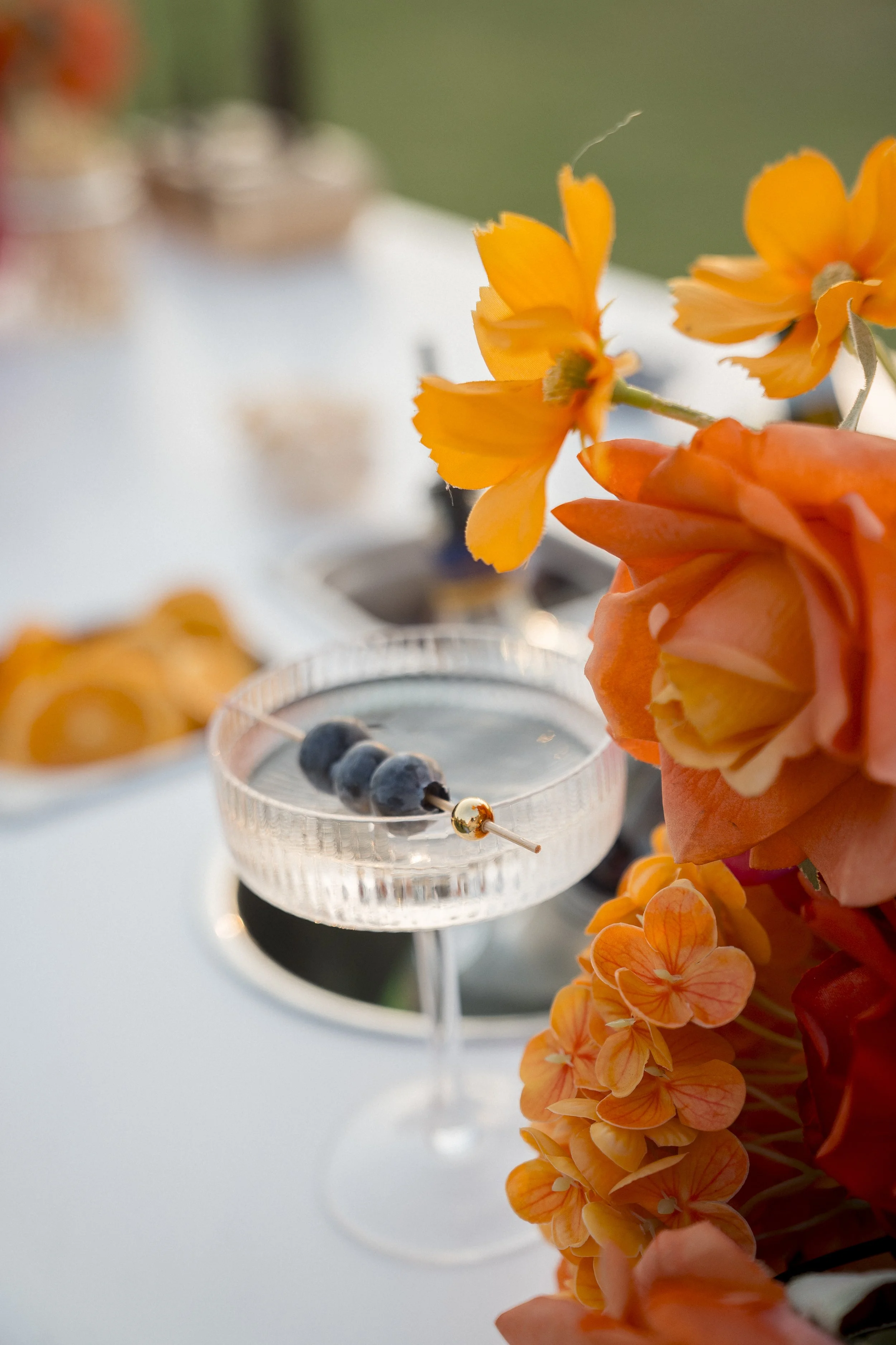 Close-up of orange flowers with a shallow depth of field, a table with food and drinks blurred in the background, and a person's hand holding one of the flowers.