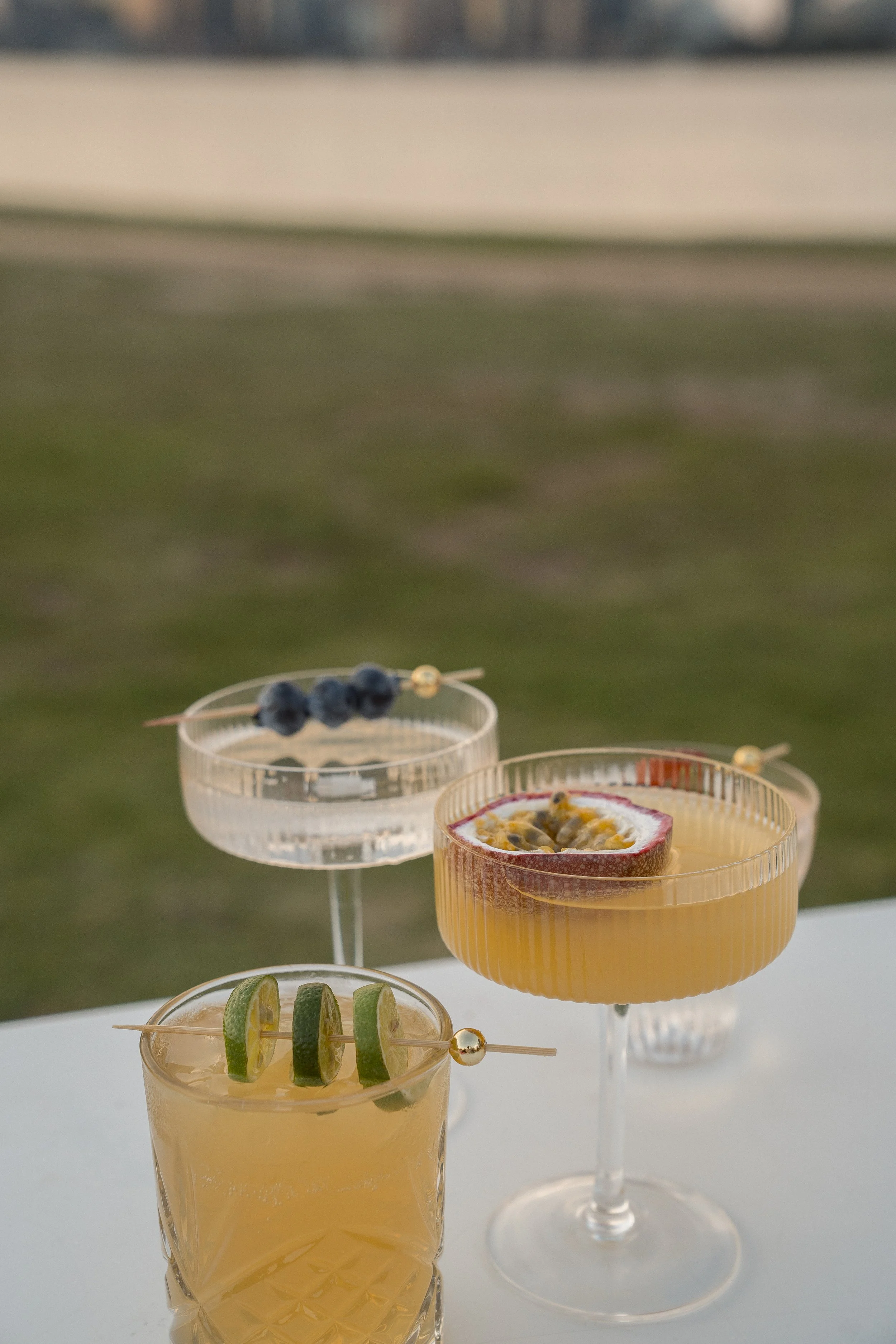 Three cocktails on a white surface with blurred outdoor background. The front glass contains a yellow drink garnished with lime slices on a skewer. The middle glass has an orange beverage with a passion fruit half as garnish. The back glass has a cle