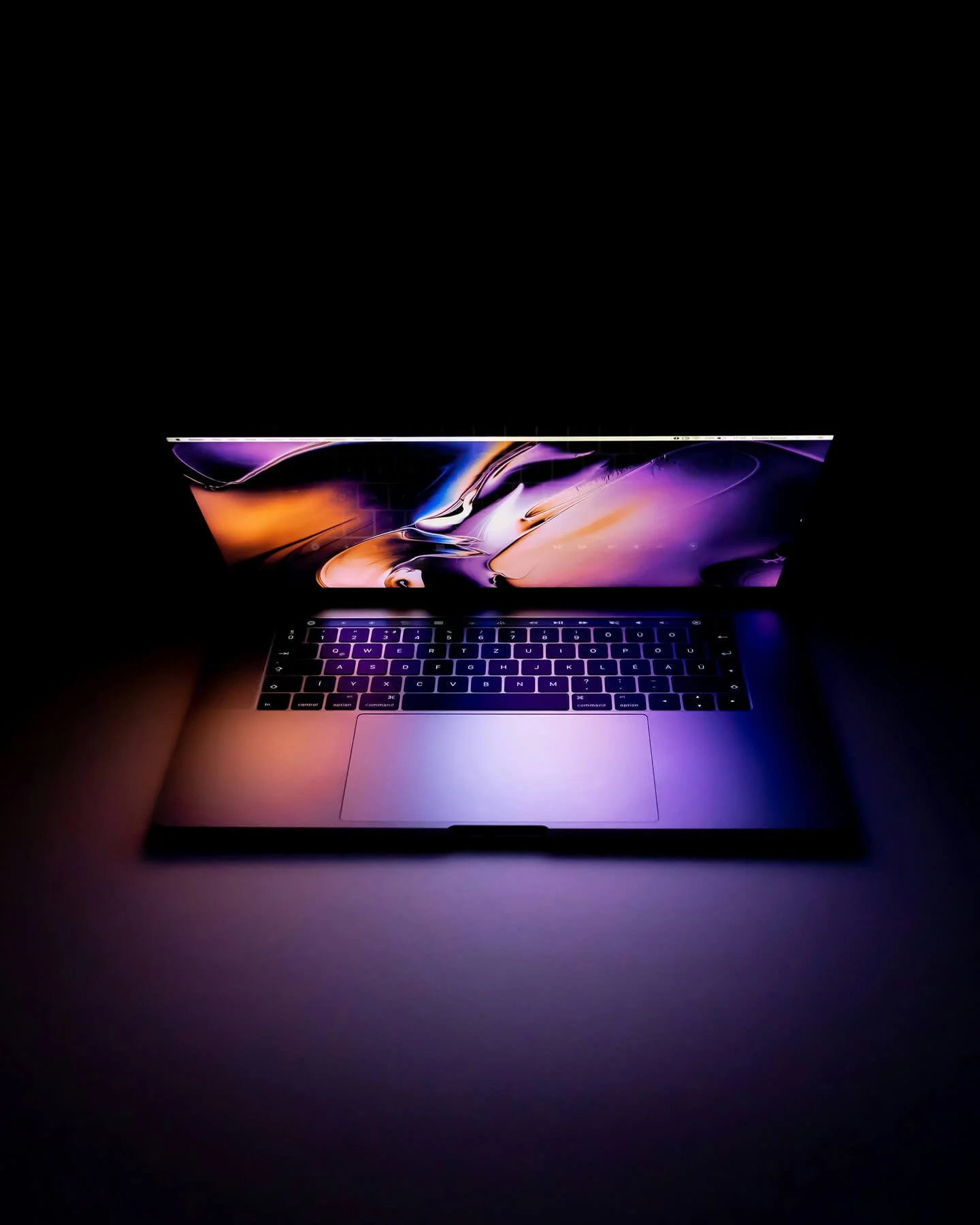 A laptop with a colourful abstract wallpaper on the screen and a backlit keyboard in a dark environment.