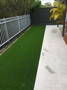 A backyard with a lawn on the left and a concrete patio on the right, separated by a narrow strip of mulch. There is a fence and some trees and shrubs in the background.