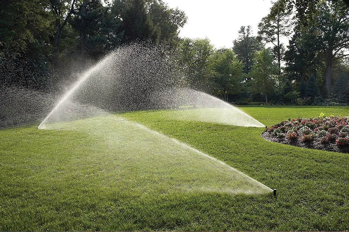 Sprinklers watering a lush green lawn with trees and flowers in the background.