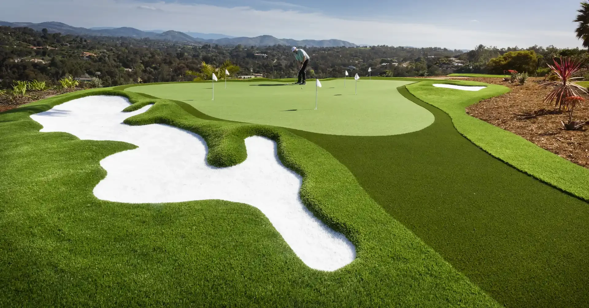 A mini golf course with a man preparing to putt a golf ball on the green, near sand bunkers and decorative plants, with a scenic hillside view in the background.