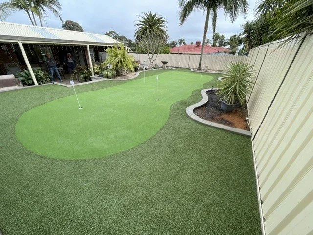 Backyard with artificial putting green, two flagsticks, tropical plants, palm trees, and a covered patio area.