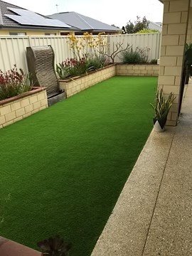 A backyard with a well-maintained artificial grass lawn, bordered by brick walls with potted plants and a garden bed with plants along the fence. There is a patio area with a concrete surface.