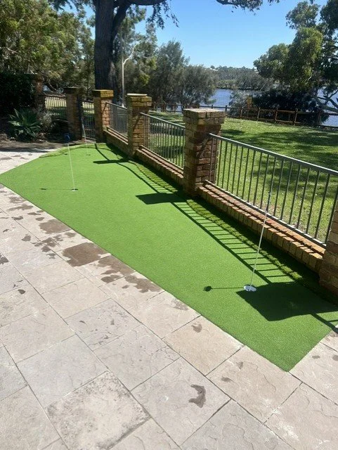 Miniature golf putting area with artificial turf and two golf holes, bordered by a brick and metal fence, overlooking a pond and trees.
