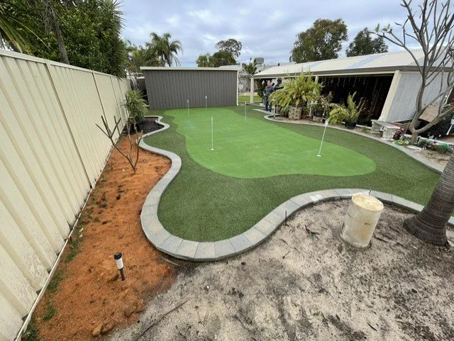 A backyard with a partially completed mini golf putting green, two holes with flags, bordered by a curved stone edging, with surrounding garden and patio area, a gray shed, and some potted plants.
