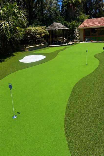 A miniature golf course featuring a putting green with a hole and flag, a sand trap, a gazebo, and a building with a red roof surrounded by trees.