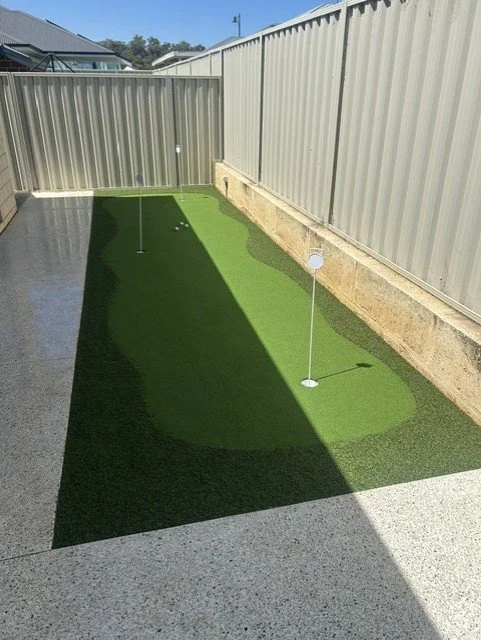 A backyard putting green with two holes and flags, surrounded by a concrete patio and high metal fence.