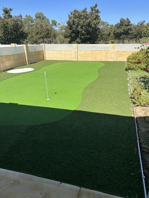 Backyard with a putting green, a sand trap, and a flagstick, enclosed by a beige brick wall, with trees and blue sky in the background.