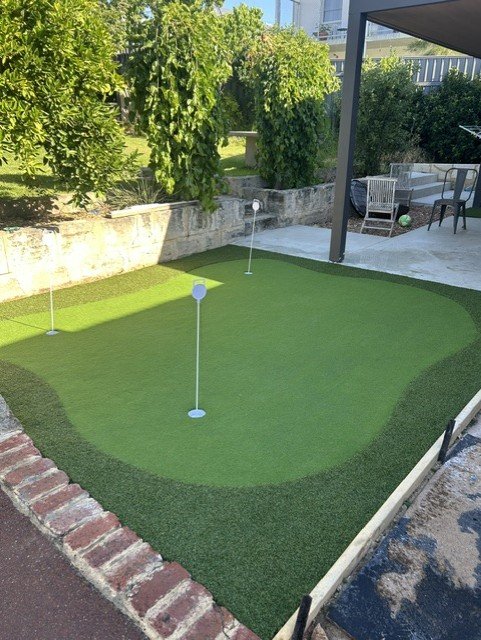Miniature golf putting green with three flags, surrounded by a brick border, in a backyard with trees and outdoor furniture.