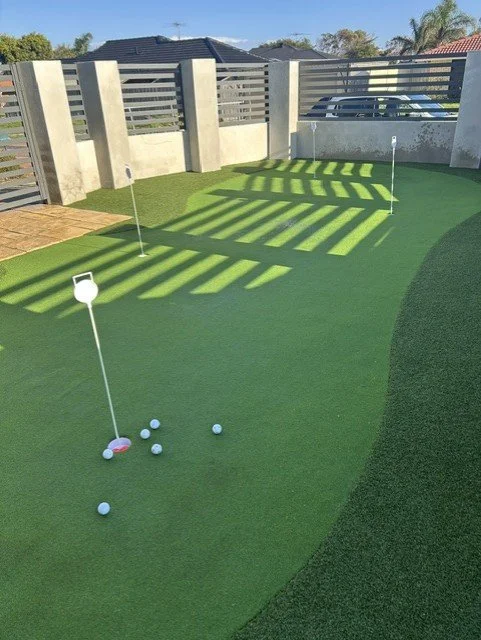 Miniature golf putting green with two holes marked by flags, several golf balls, and a small pink tee. Shadows from a slatted fence create striped patterns on the grass.