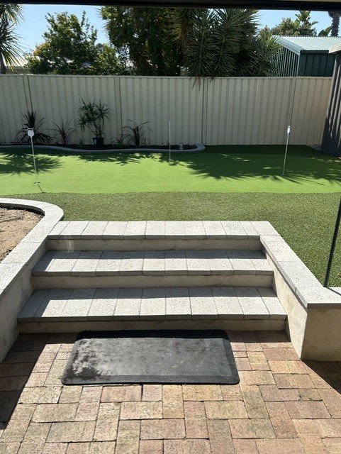 Backyard with artificial grass, small garden beds, and a boundary fence, viewed from a patio with steps and a black doormat.