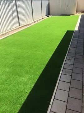 Artificial grass lawn next to a paved walkway, enclosed by a metal fence, with sunlight casting shadows.