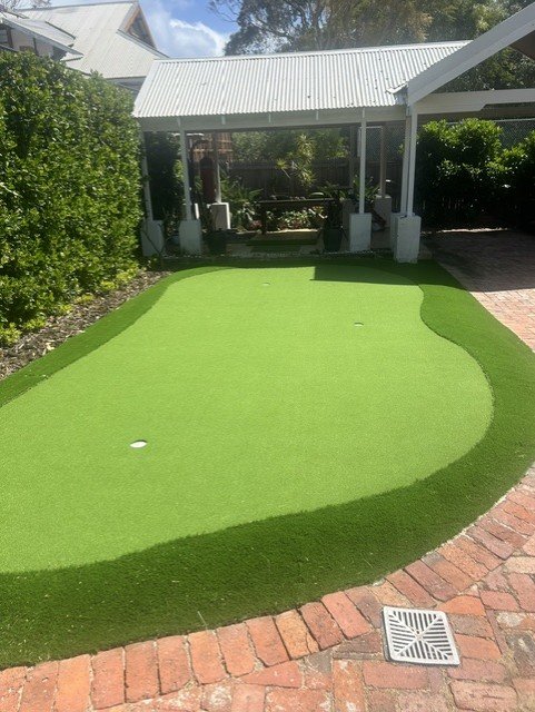 A backyard putting green with a hole, surrounded by red brick pavers, next to a screened porch and green shrubbery on a sunny day.