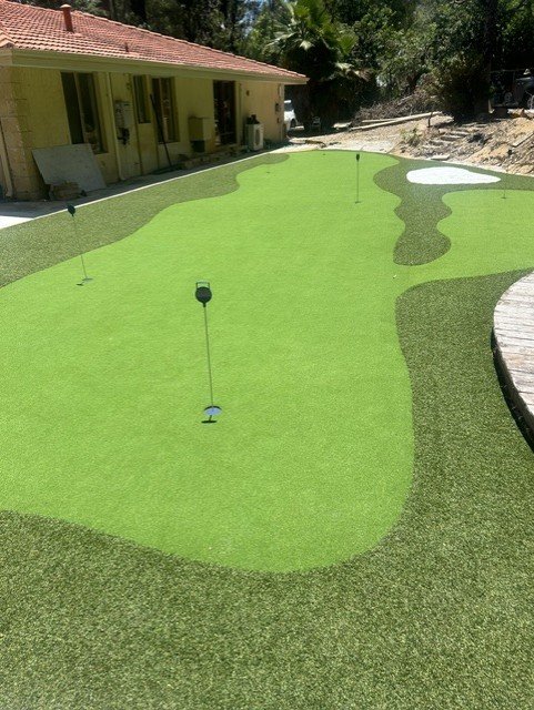 Miniature golf course with putting green, sand trap, and flags, adjacent to a house with a red-tiled roof.