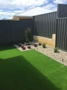 Backyard with artificial green grass, a small landscaped garden, and a dark metal privacy fence.