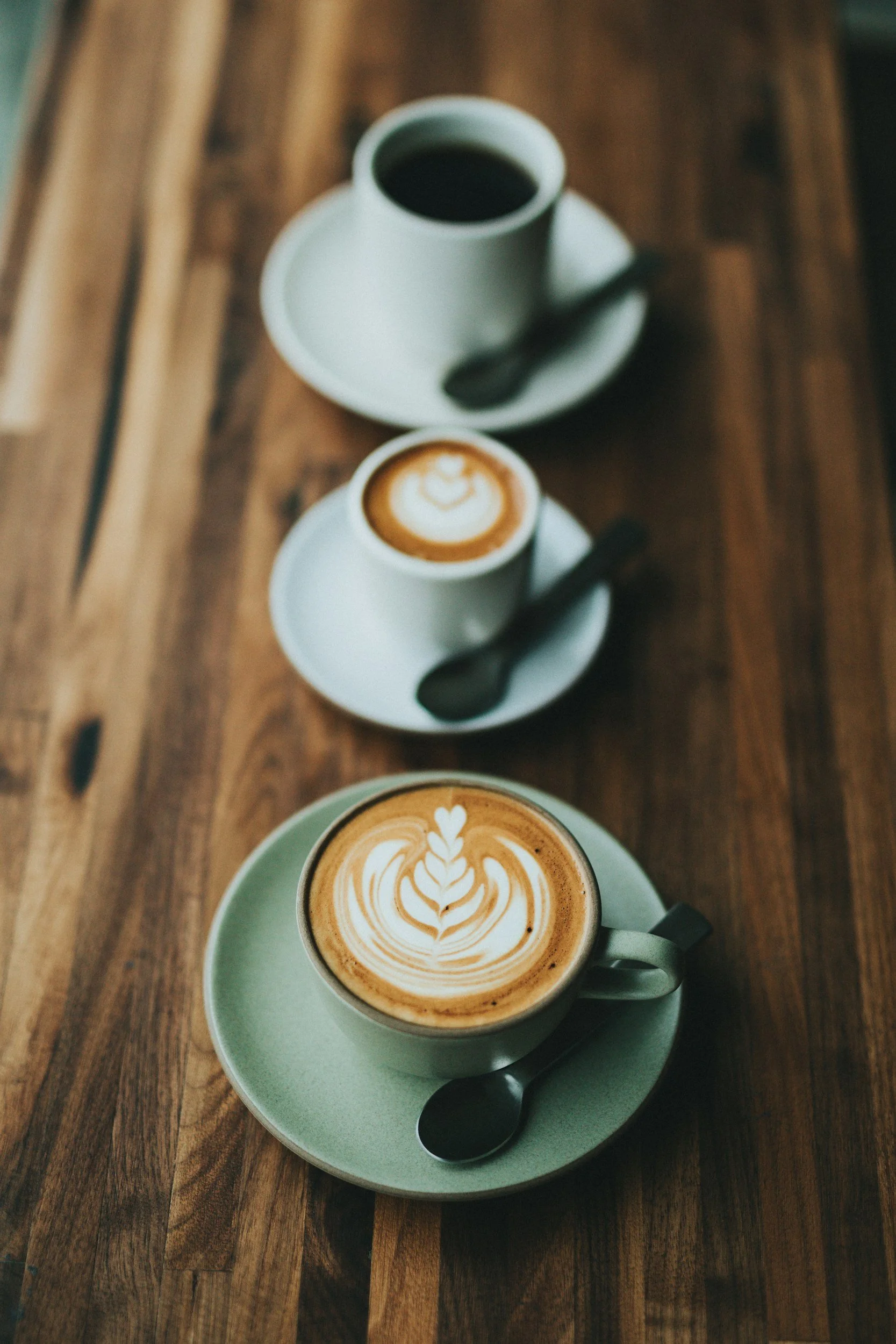 A top view of a latte with latte art in a gray cup on a matching saucer, along with two other cups of coffee with dark beverages on a wooden table.