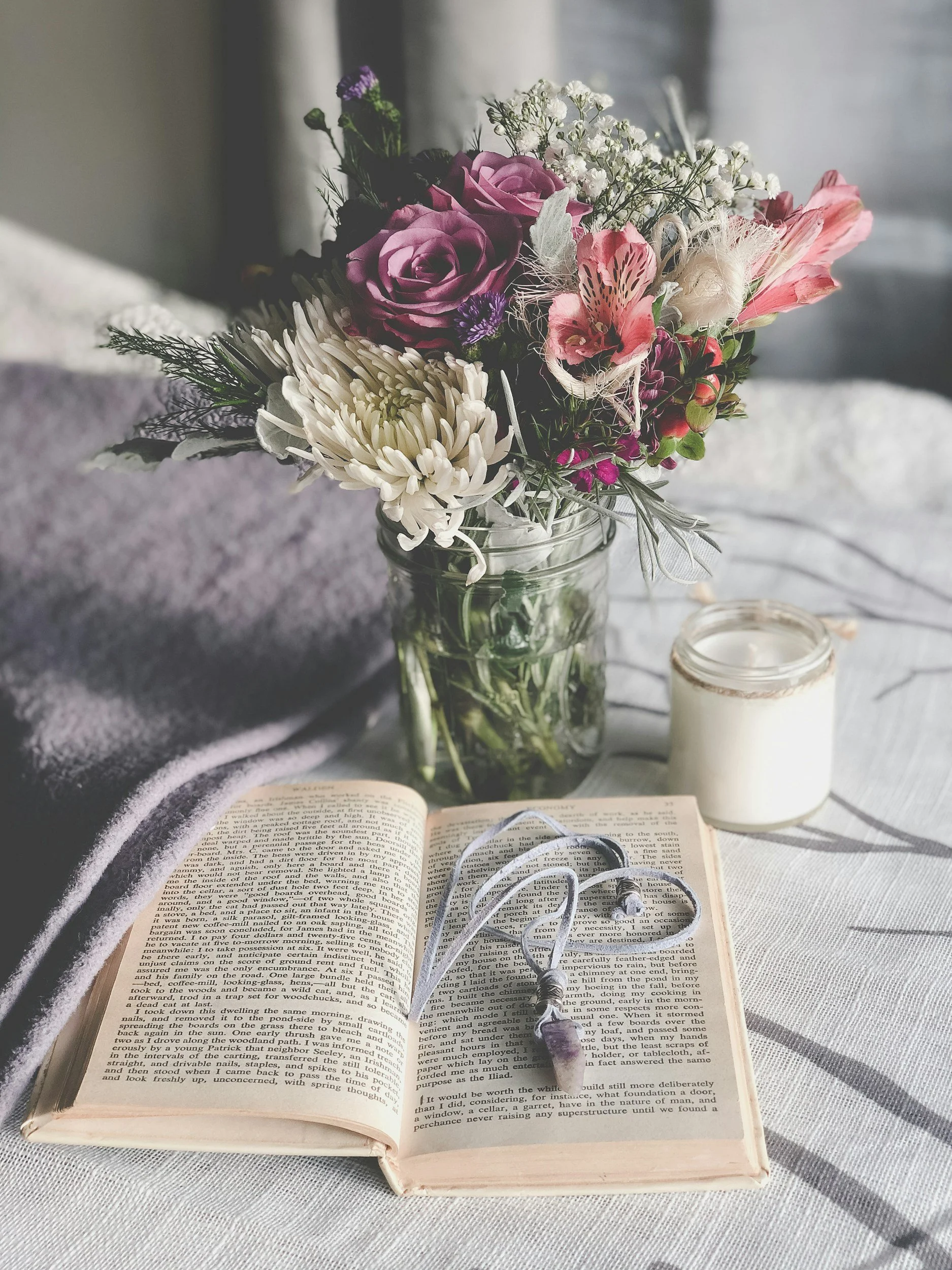 Open book with black and white photograph, clear text, a purple and white flower bouquet in a jar, candles, on a gray fabric surface with sunlight and shadows.