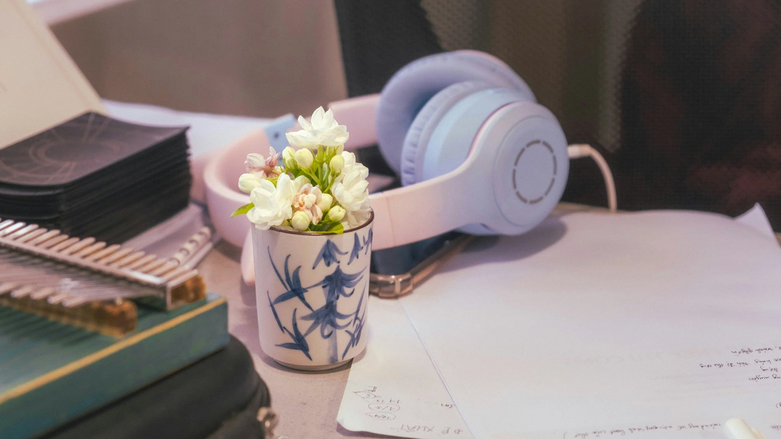 A desk with a small vase of white flowers, a pair of white headphones, a stack of black cards, and some papers with notes.