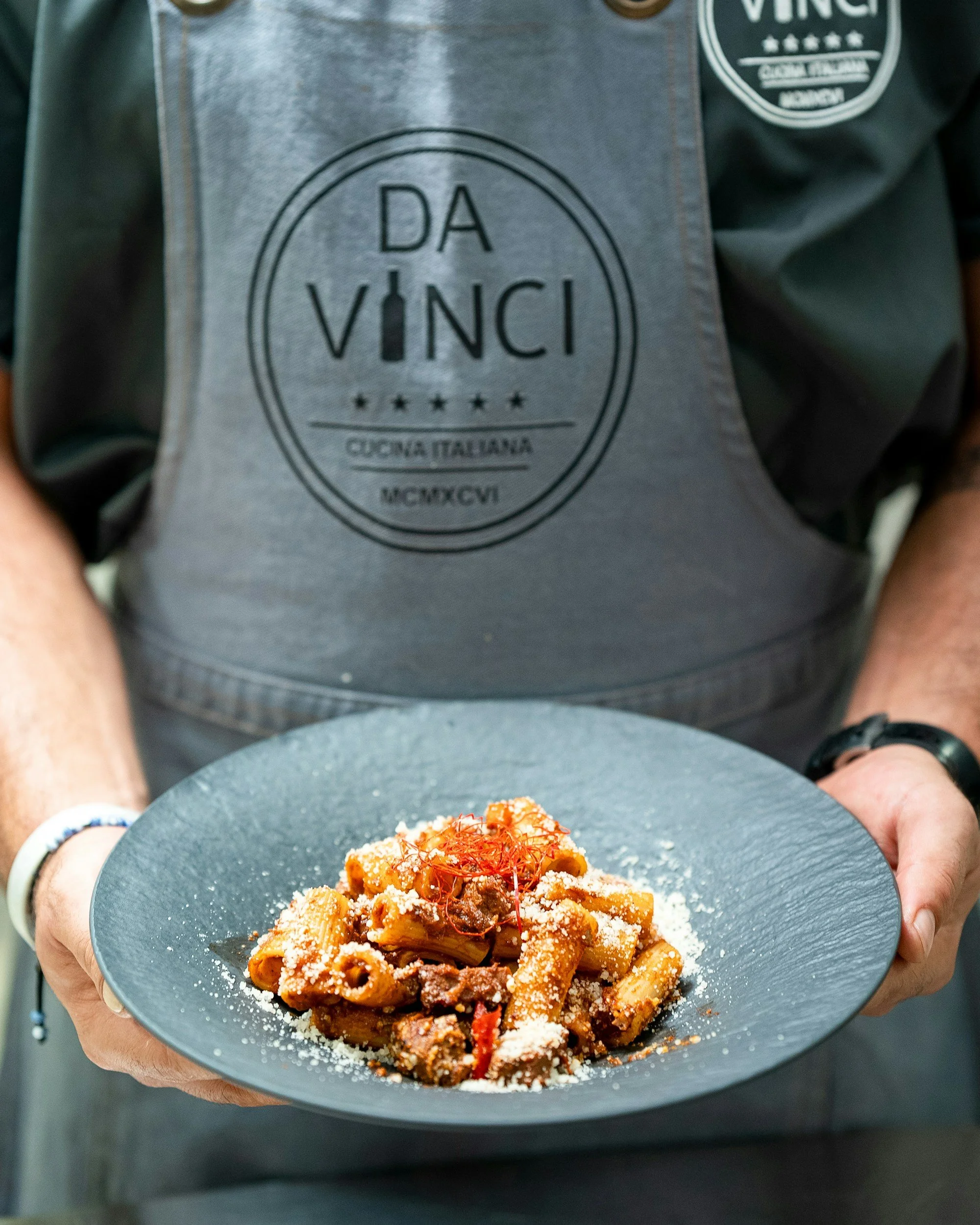 Persona che tiene un piatto di pasta con sugo, formaggio grattugiato e guarniture, indossa un grembiule con il logo 'Da Vinci' e un orologio al polso.