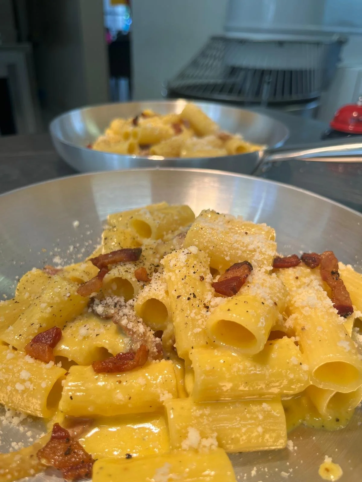 Pasta corta ripiena con pancetta e formaggio, servita in una ciotola di acciaio inossidabile. Sullo sfondo, ci sono due ciotole di pasta, in una delle quali si vedono pezzetti di pancetta e formaggio.