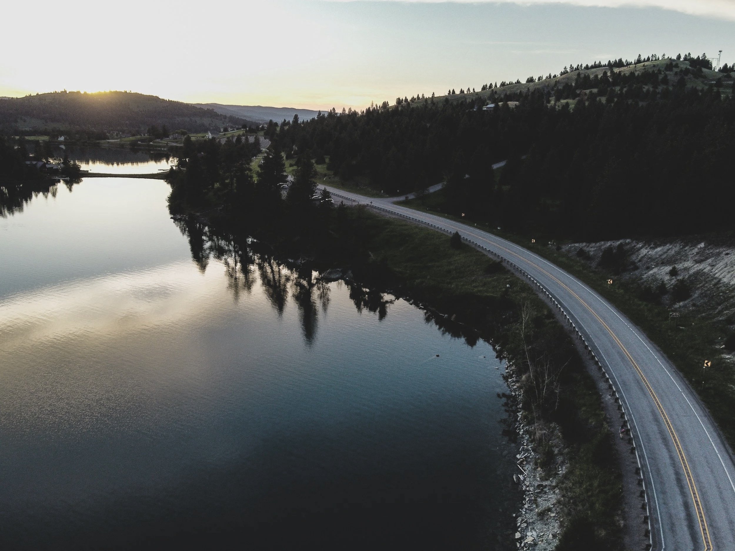 A scenic view of a lake with a winding road on the side, surrounded by forested hills during sunset or sunrise.