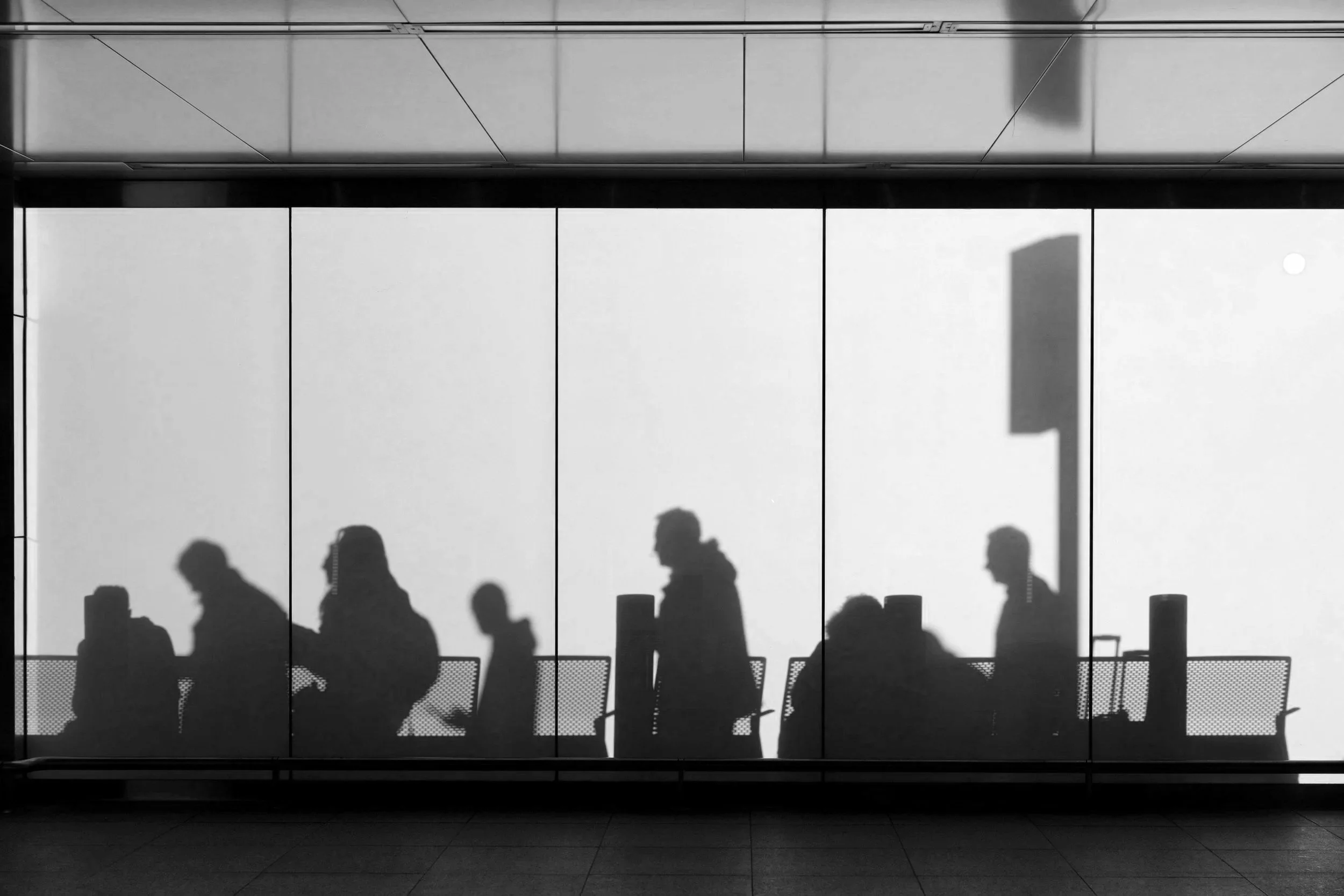 Shadowed silhouettes of people walking past a large window or glass wall with a reflective surface and blinds at the top.
