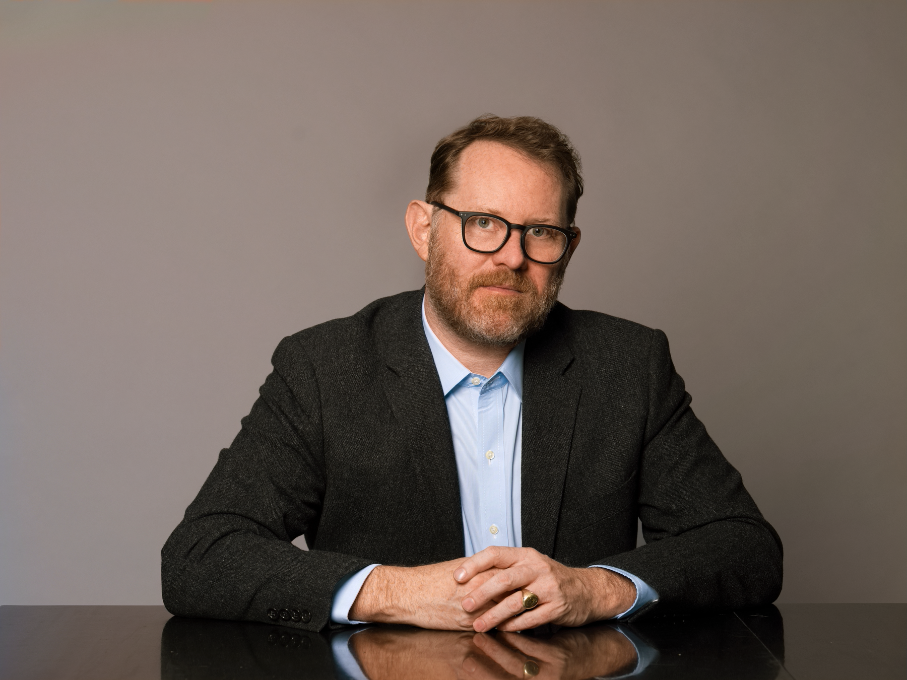 A man with glasses and a beard, wearing a navy blazer over a light blue shirt, sitting at a dark table against a plain gray background, looking directly at the camera with his hands clasped.