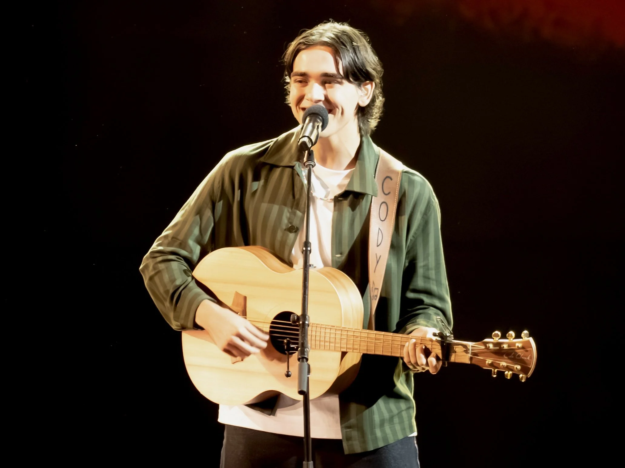 Young man with dark hair playing an acoustic guitar on stage, singing into a microphone, wearing a plaid shirt over a white t-shirt, with a black background and red stage lighting.