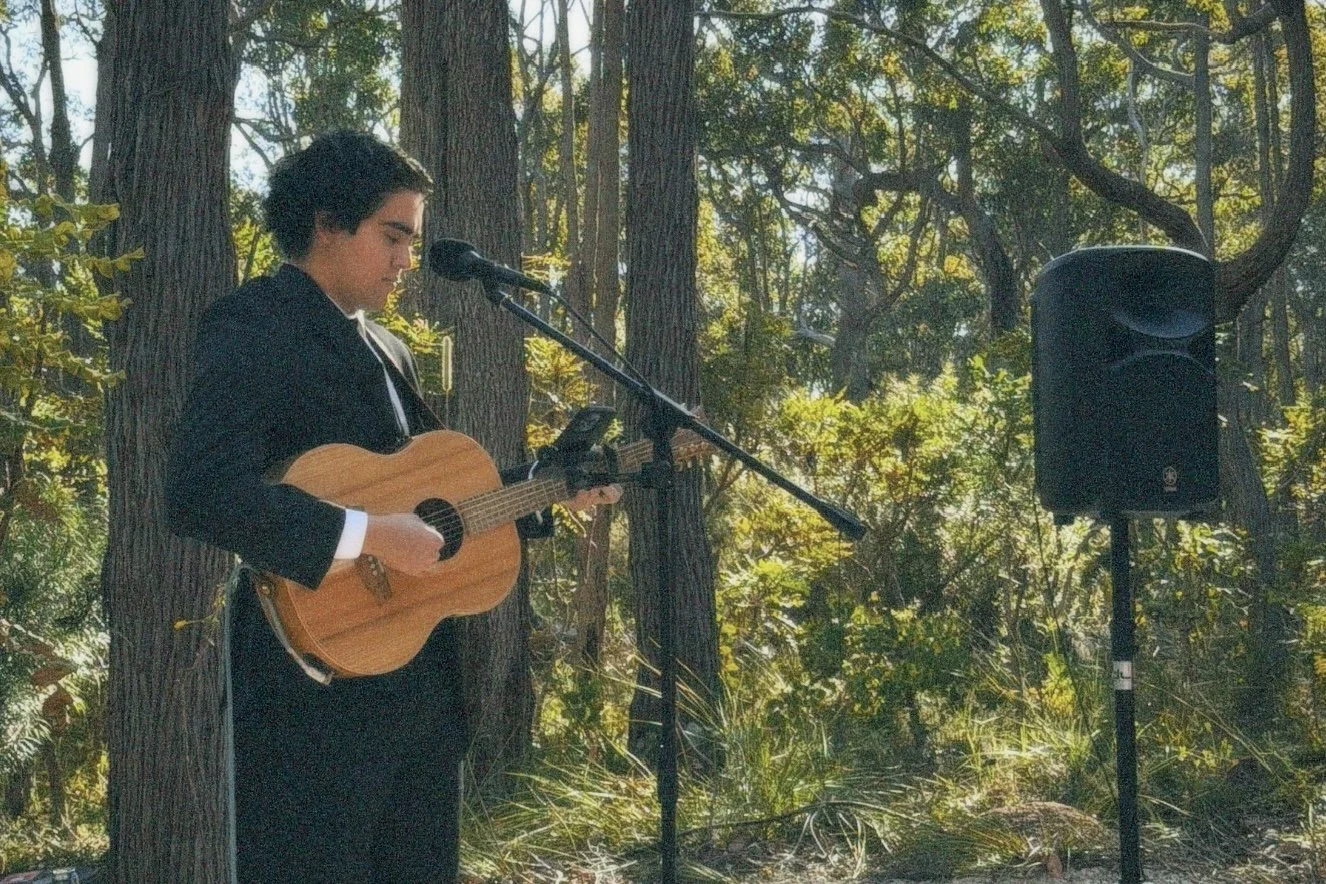 A person in a black jacket and white shirt playing an acoustic guitar at an outdoor setting among trees, with a microphone and speaker nearby.