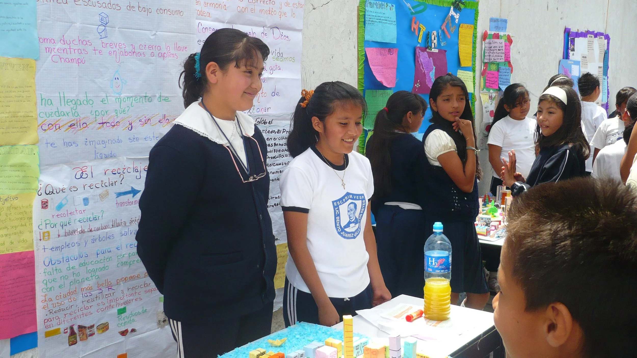Grupo de niños en una feria escolar presentando un proyecto relacionado con el cuidado del medio ambiente, con carteles coloridos y figuras en la mesa.