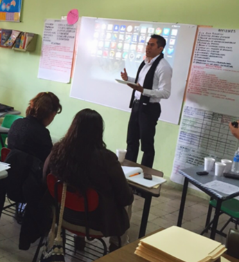 Un profesor de pie frente a una pizarra digital en un aula, explicando a tres estudiantes sentados