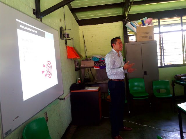 Un hombre de pie frente a una pizarra en un aula, dando una presentación o clase, en un entorno con ventanas y muebles escolares.