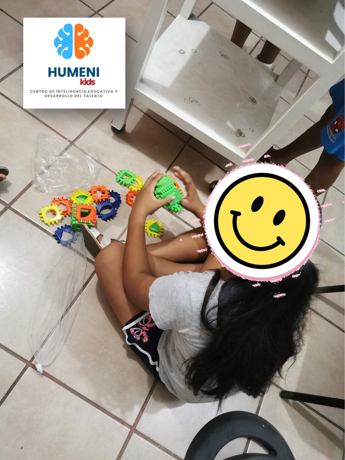 Niña jugando con piezas educativas de colores en el piso, con una silla blanca y una mesa blanca al fondo en una habitación con piso de baldosas.