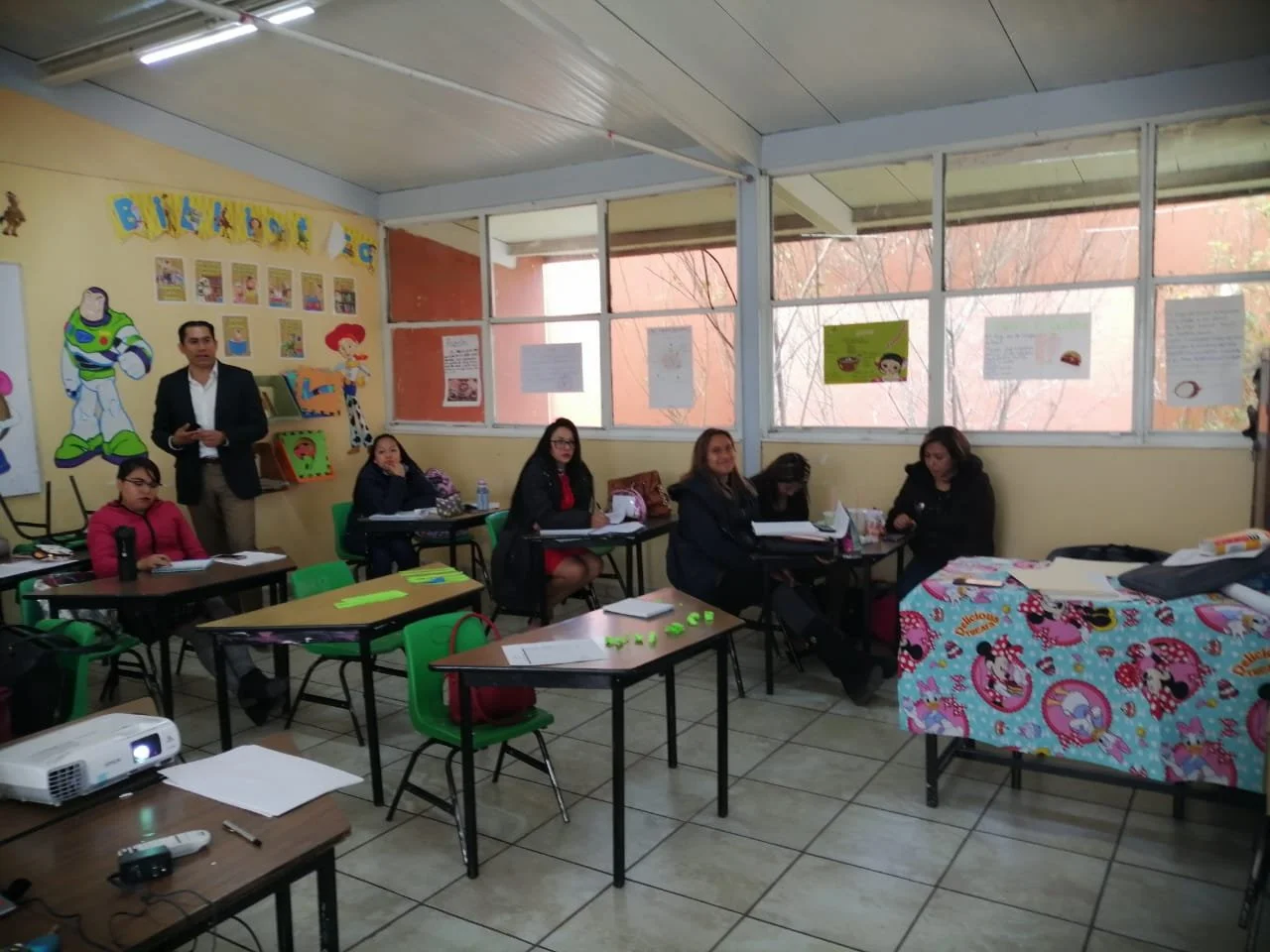 Grupo de personas en un aula, algunos sentados en sillas verdes y otros de pie, con paredes decoradas con personajes de dibujos animados y carteles, y ventanas grandes que dejan ver árboles afuera.