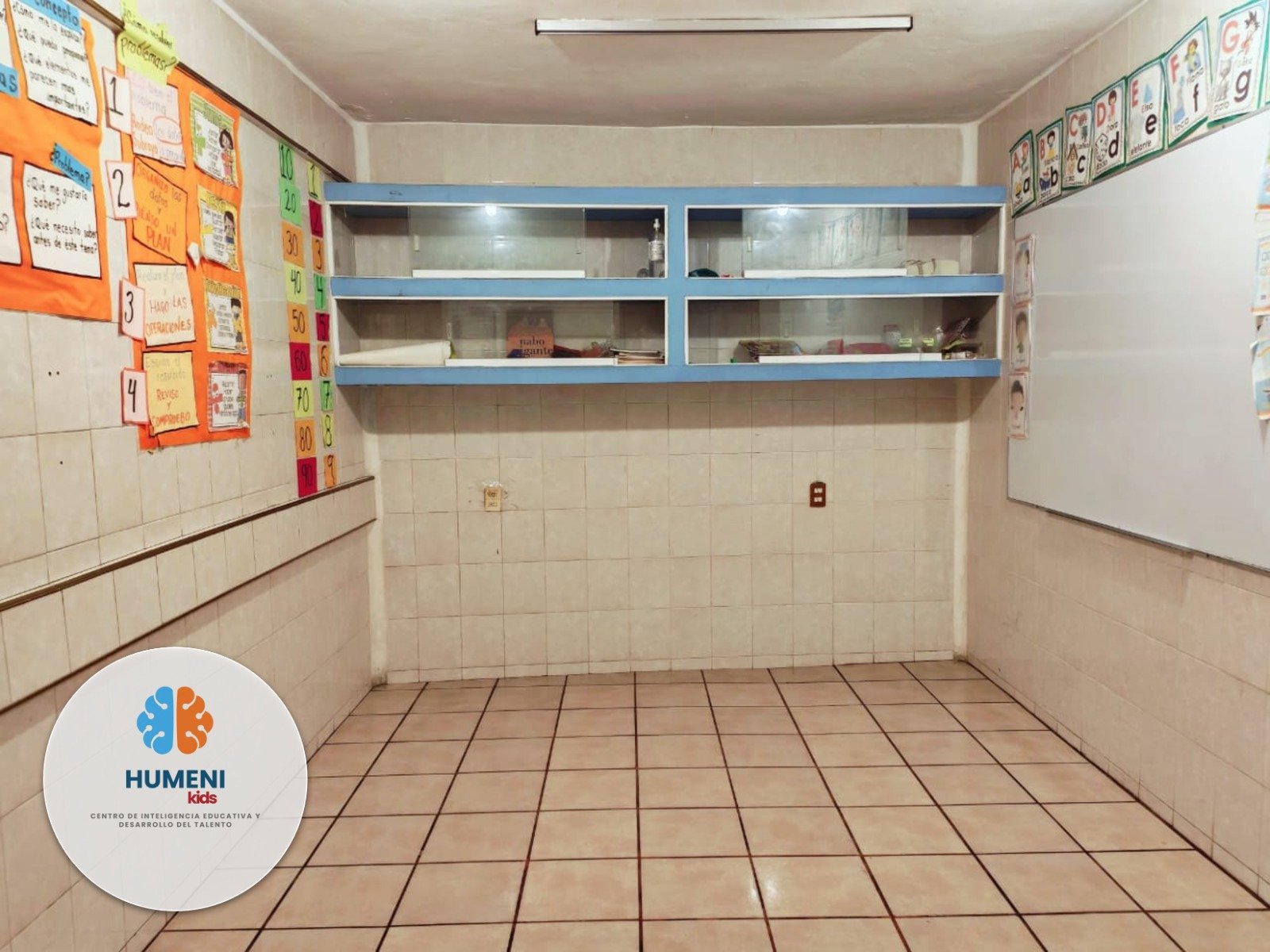 Un aula escolar con estantería de vidrio en la pared y cartel con actividades escolares en la pared a la izquierda.