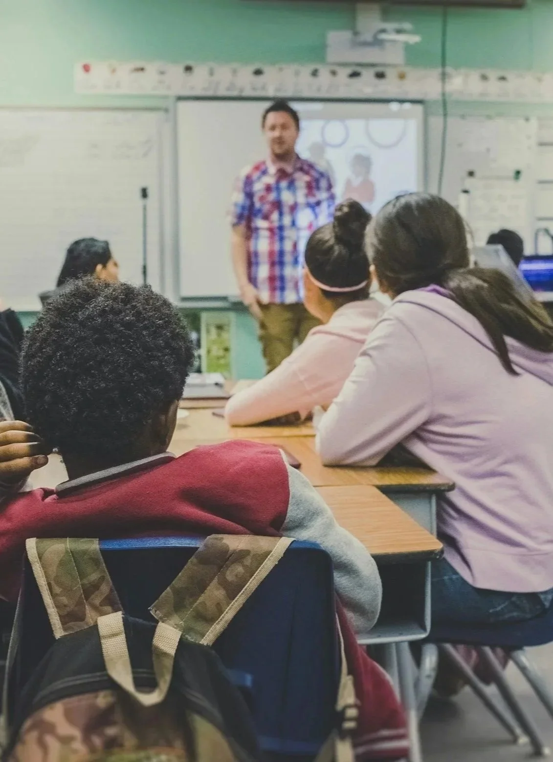 Un profesor dando una clase a estudiantes en un salón, con una pizarra y una pantalla en el fondo.