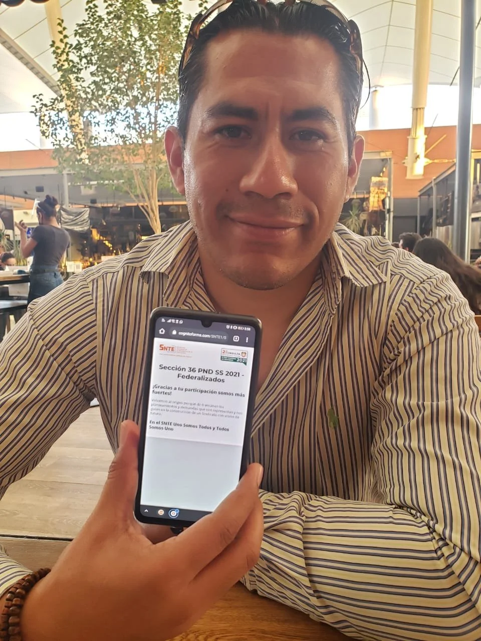 Hombre sonriendo y mostrando una pantalla de teléfono móvil con texto sobre educación, sentado en un restaurante o cafetería con fondo de árboles y estructura de techo moderna.