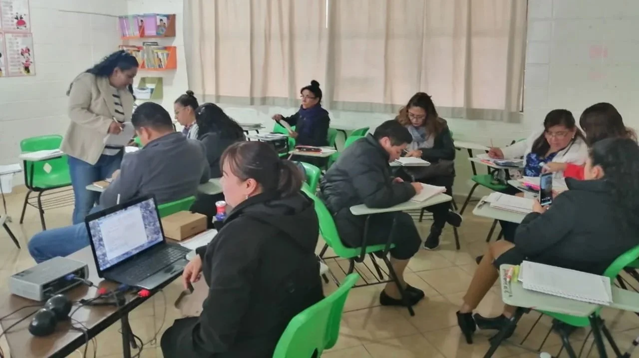 Grupo de personas en un salón de clases o reunión, sentados en escritorios con computadoras, libros y cuadernos, algunos conversando y otros trabajando.