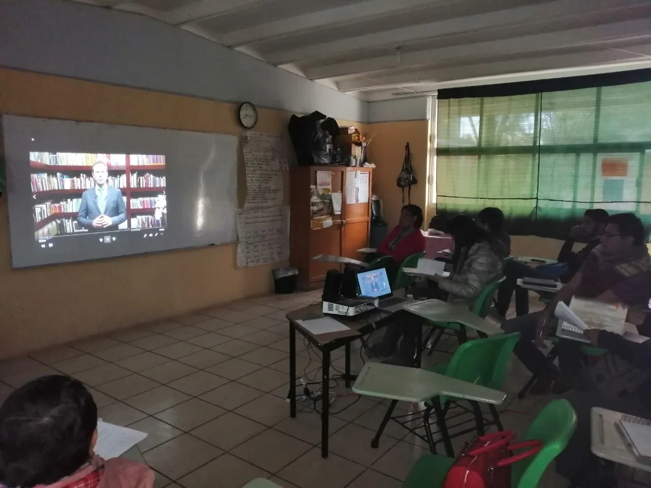 Clase en salón con estudiantes viendo una videollamada en una pantalla y tomando notas.