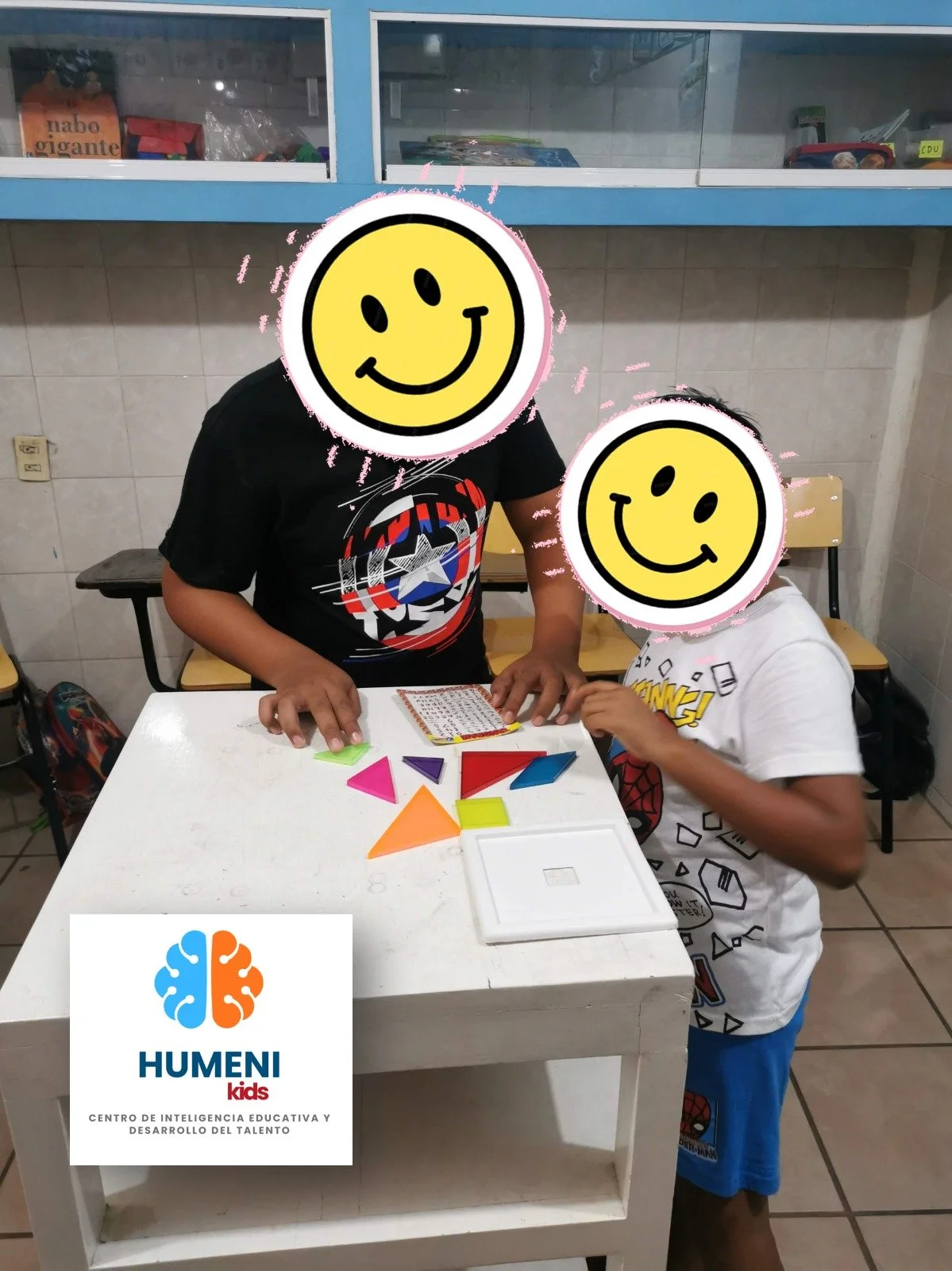 Dos niños en una mesa jugando con formas geométricas de colores en un aula con muebles y estantes despoblados.